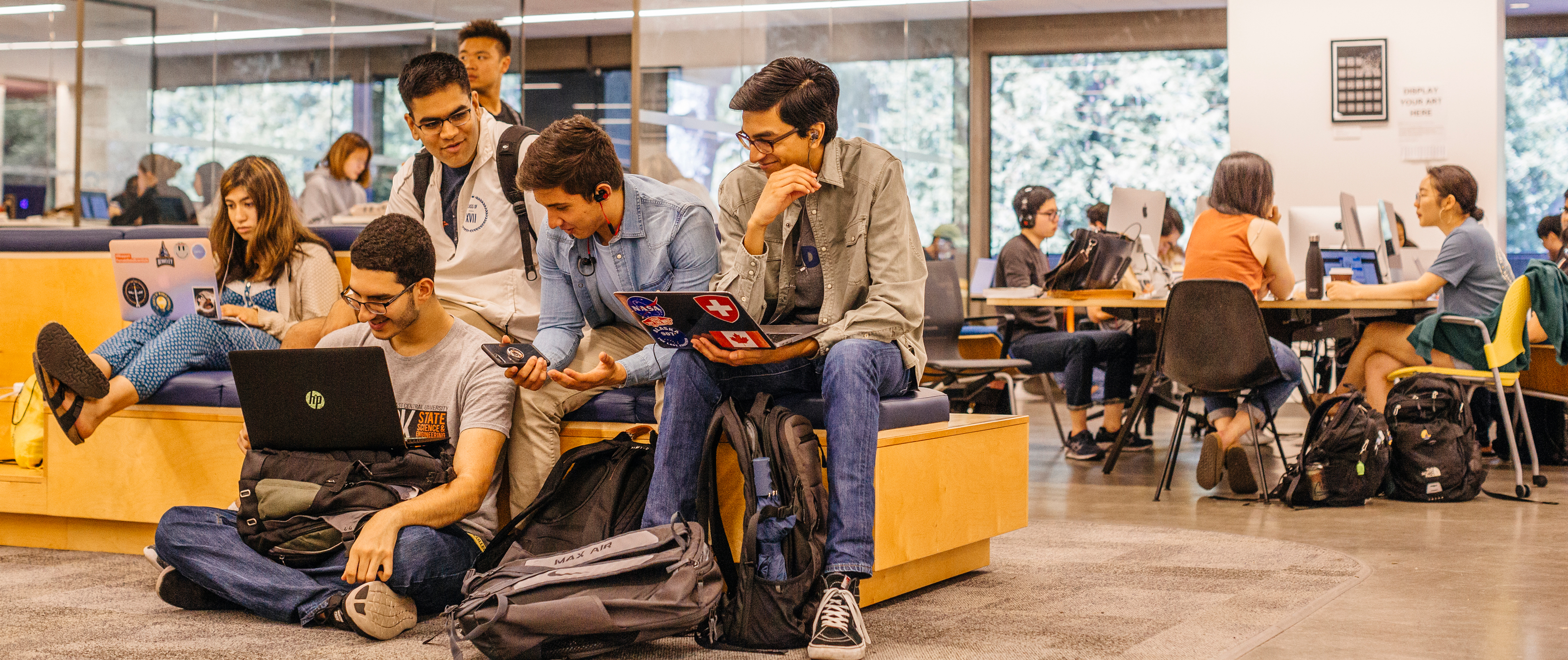 A group of college students work together in a modern campus study area. Some are sitting on benches and the floor with laptops and backpacks, while others collaborate at tables with desktop computers in the background.