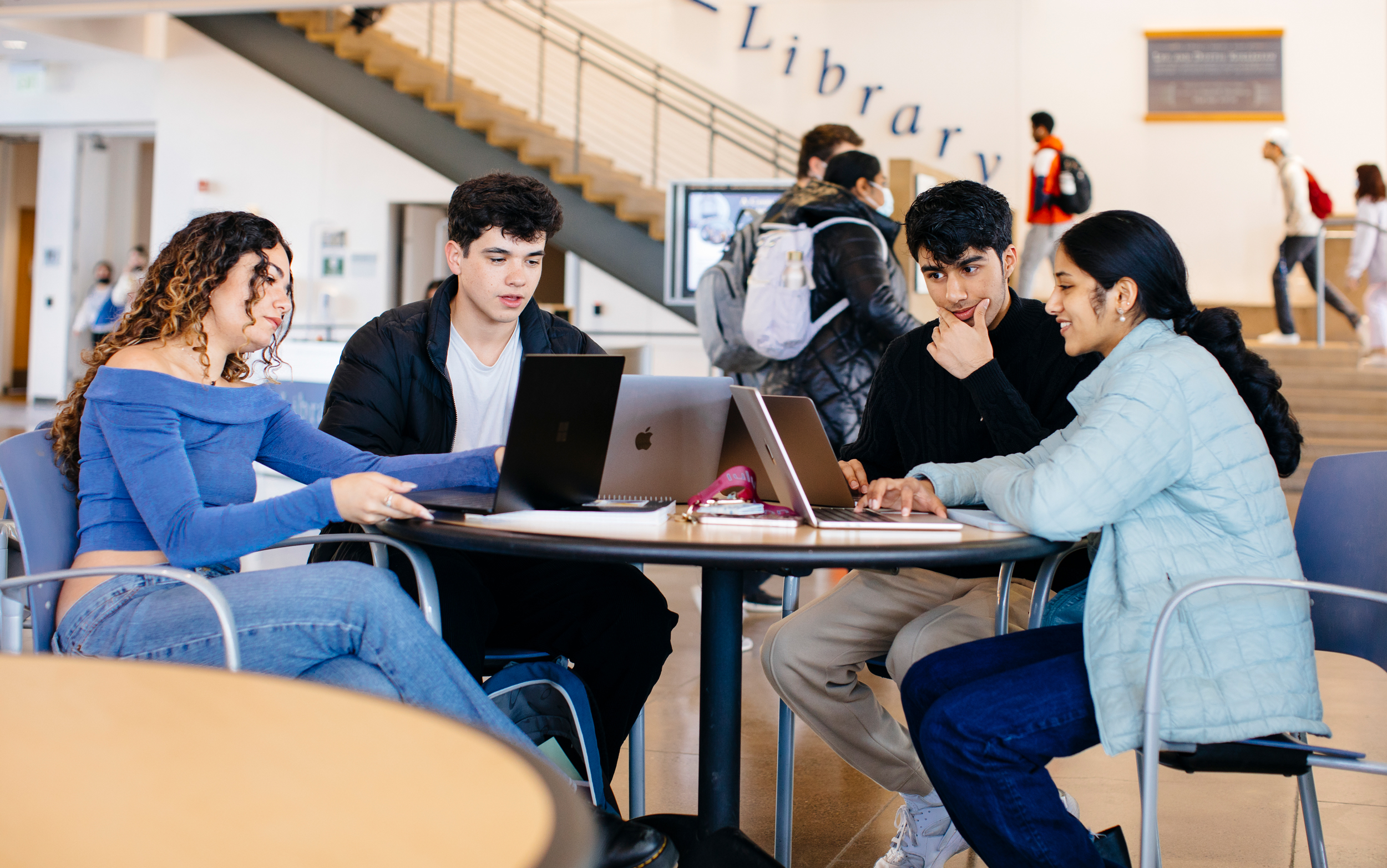 A group of students studying together at a table in a busy campus space, with a sign that says "Library" in the background. 