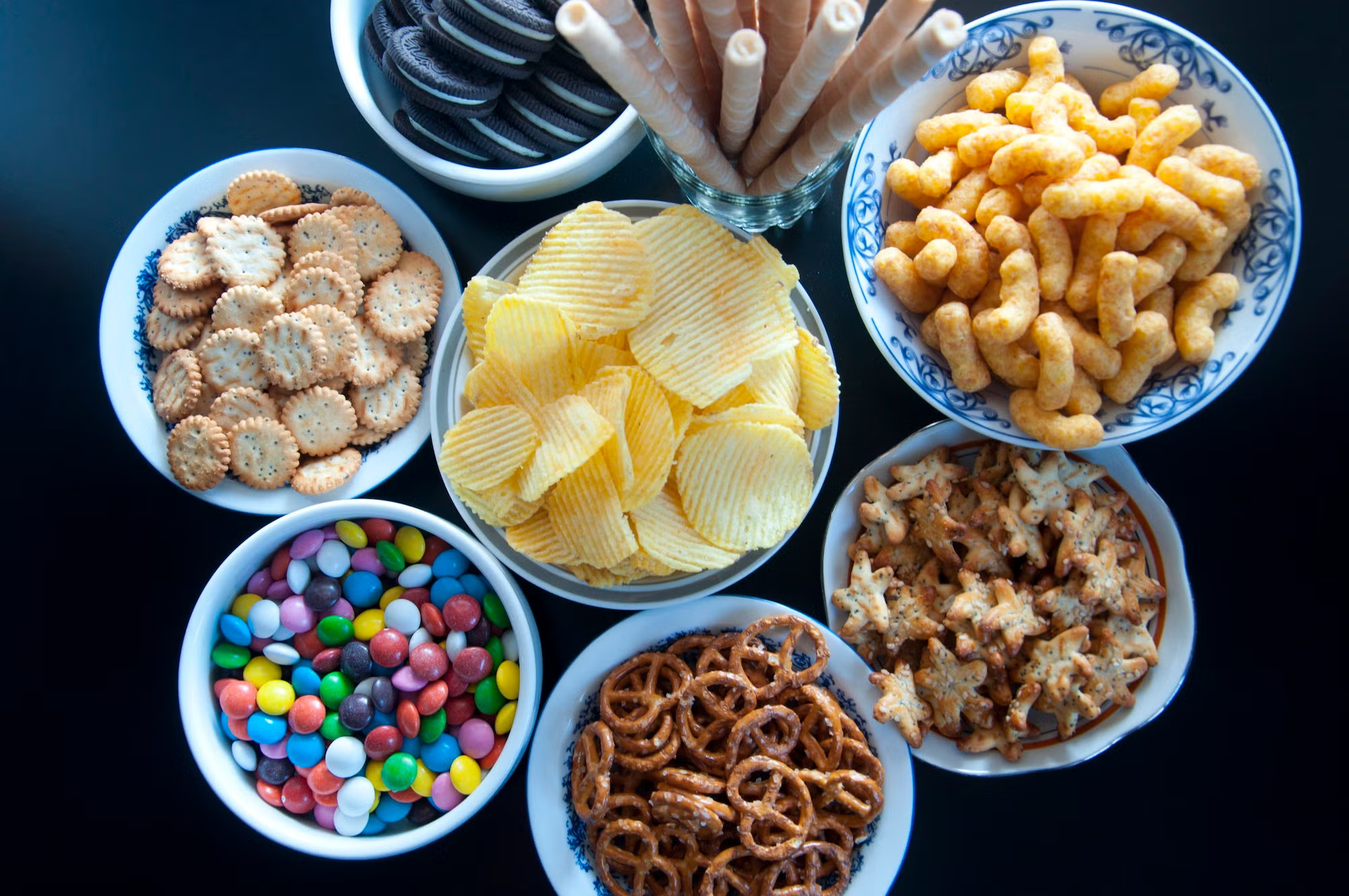 8 small bowls and a glass full of chips, pretzels, cookies and other processed foods