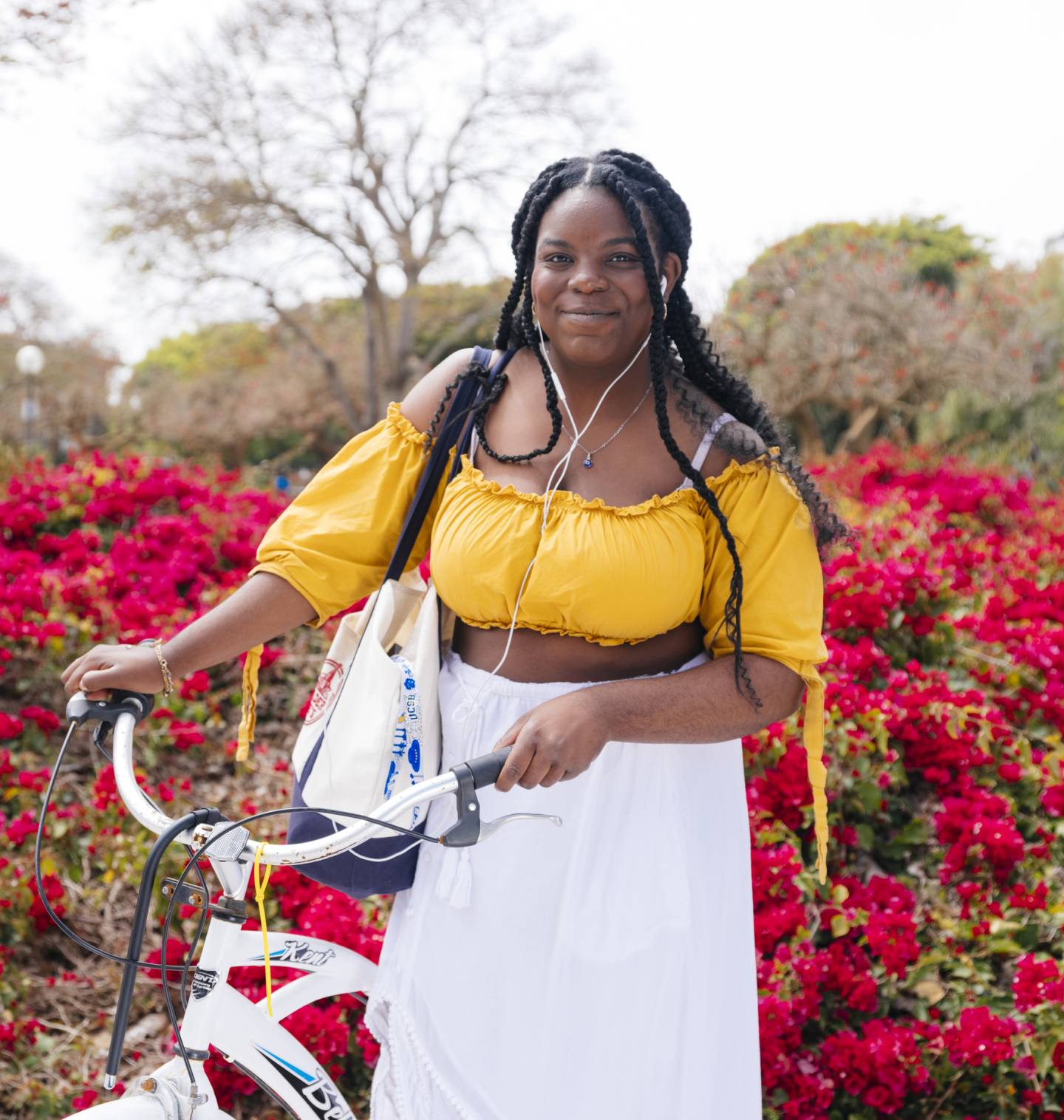 A woman on campus wearing a yellow top and white skirt stands beside a bicycle, smiling in a sunny outdoor setting.