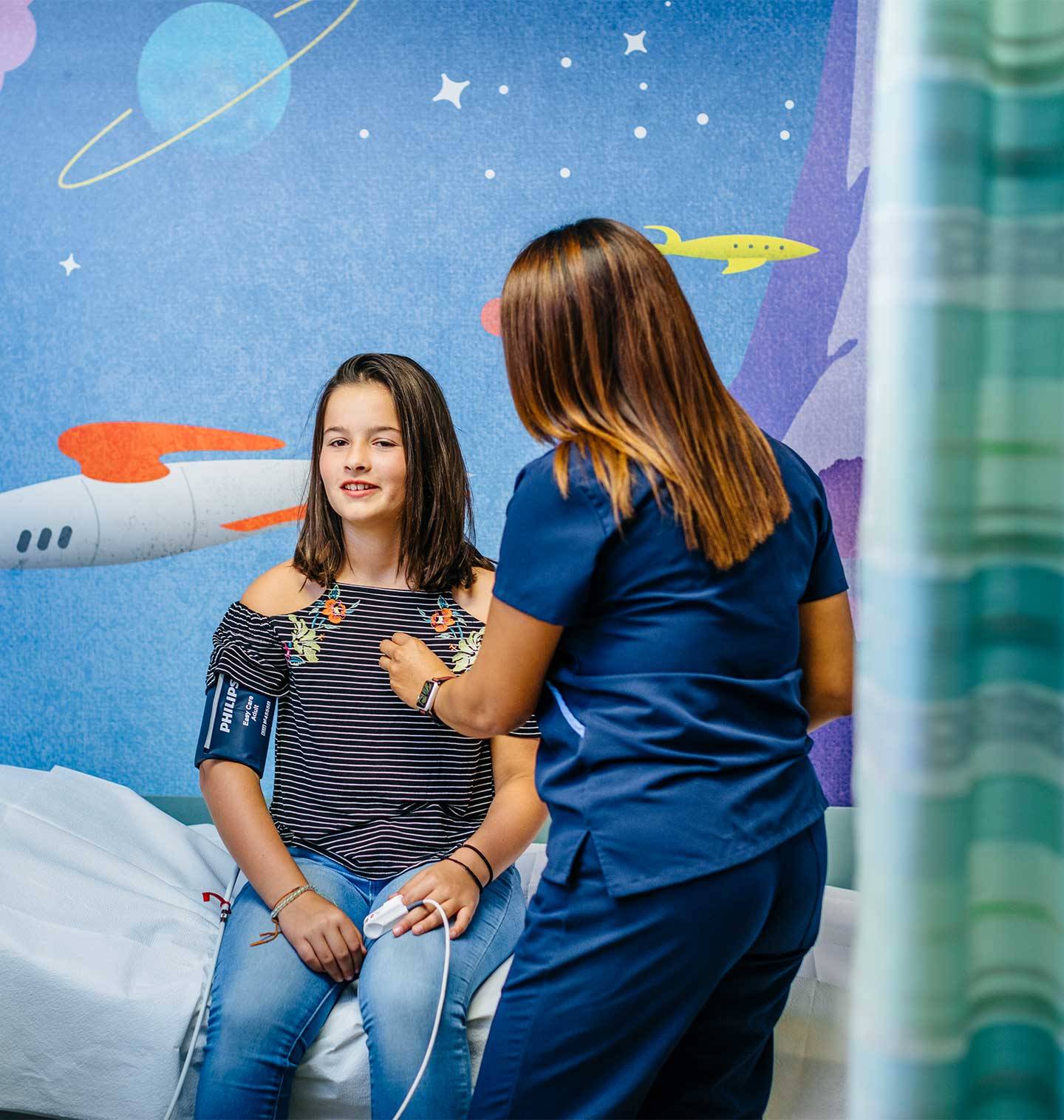 A nurse listens to the heartbeat of a young teen sitting up on a bed in a room with colorful spaceship wallpaper