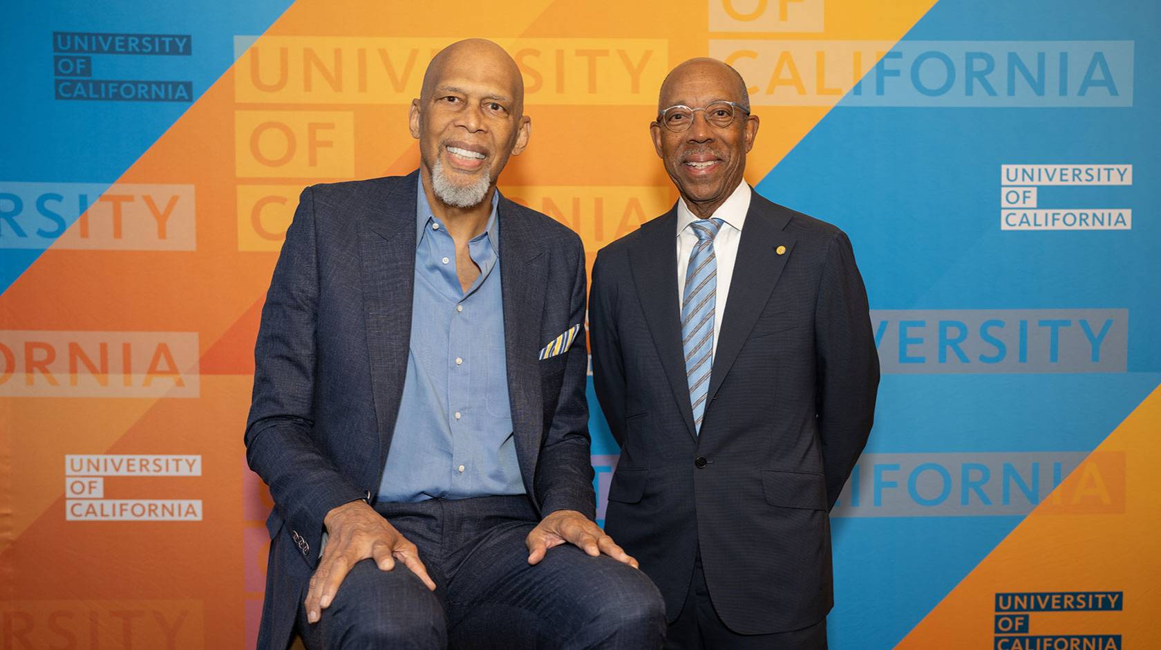 Kareem Abdul-Jabbar and UC President Michael Drake, M.D., smile for a portrait, standing in front of a University of California-branded backdrop