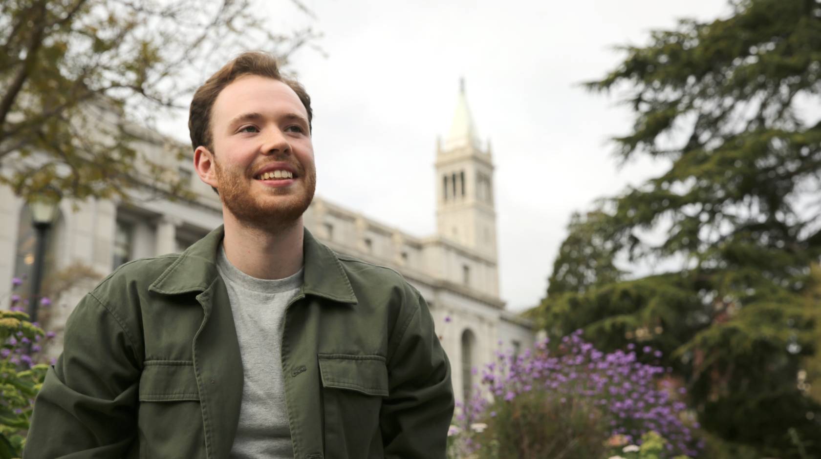 a young man wearing a green jacket puts his hands in his pockets and smiles in front of the Campanile