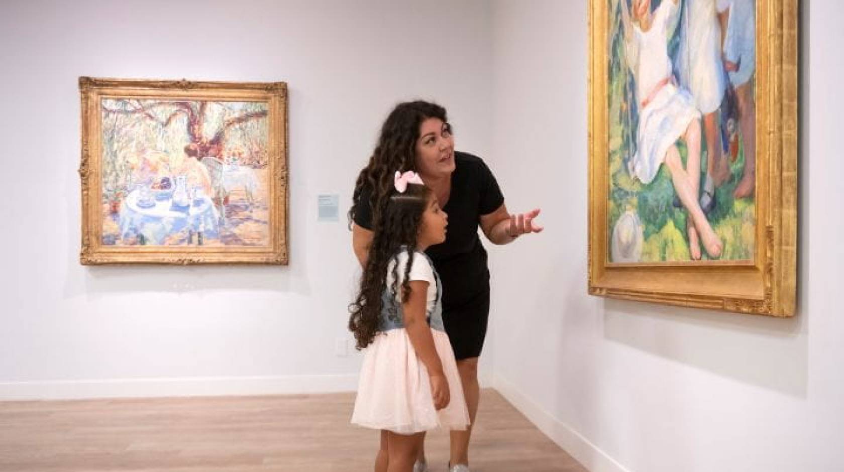 A mother and her young daughter talking about a painting they are looking in a museum gallery.