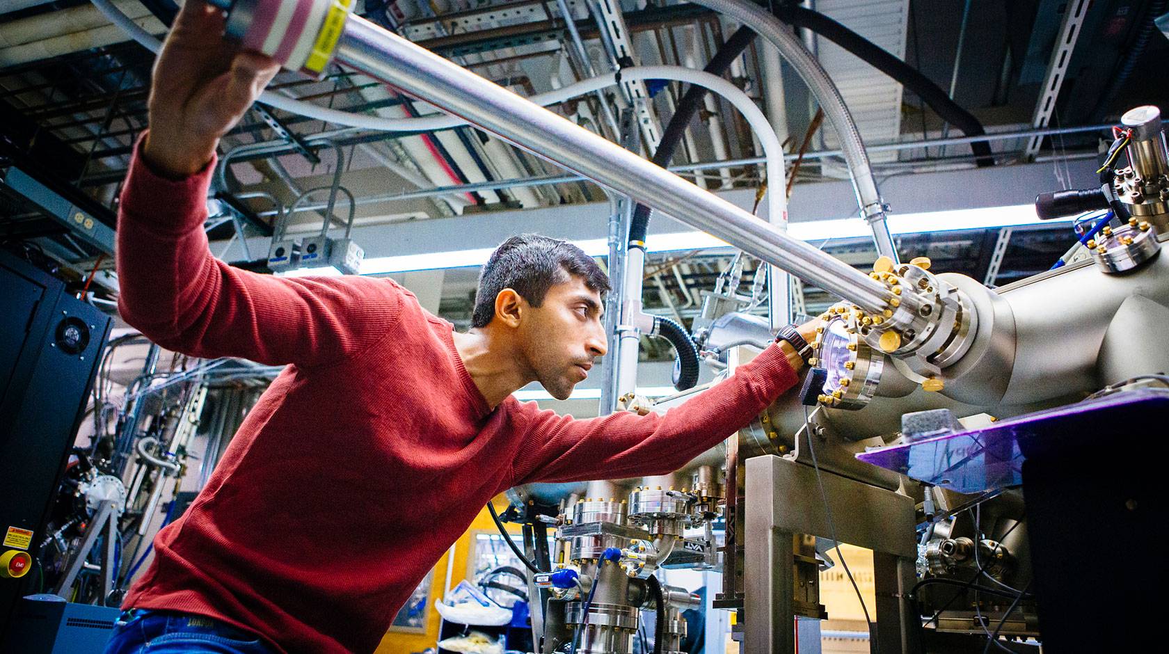 A grad student focuses intently as he slides a three-foot-long steel bar into a large apparatus made of tubes and wires, in a lab full of similar such apparatuses. 