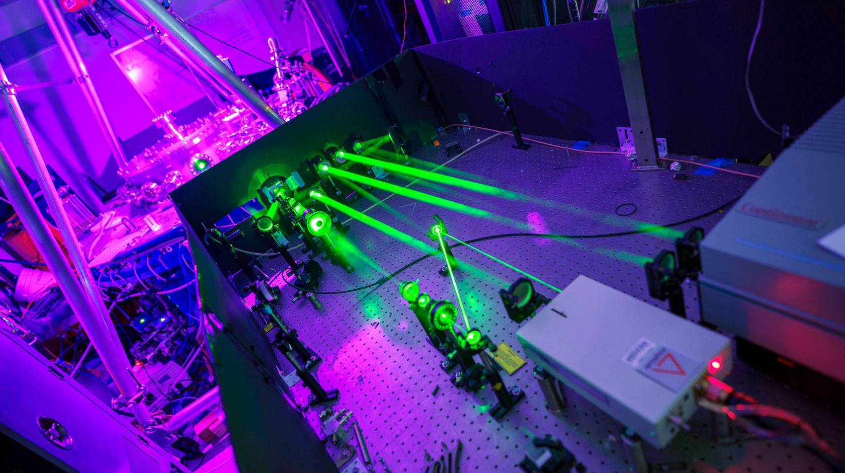 A laboratory setup with multiple bright green laser beams projected across an optical table lined with mirrors and instruments, illuminated by purple and blue ambient light, showing advanced experimental physics equipment.