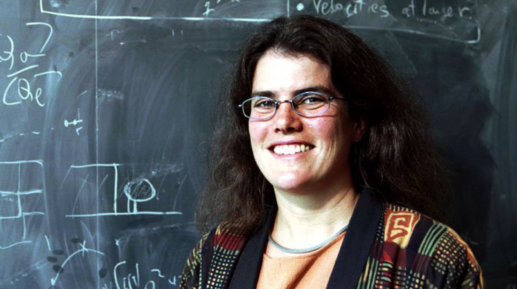 A woman wearing glasses standing in front of a chalkboard covered in scientific notations
