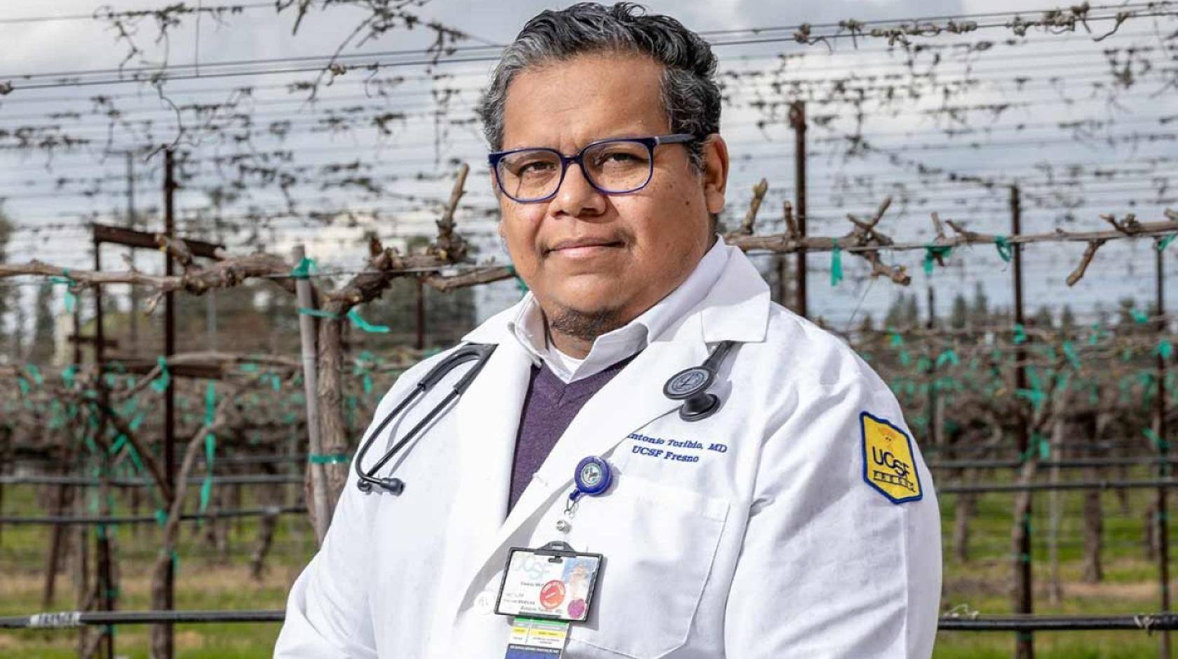 Doctor with dark gray hair and a medical coat stands in front of a vineyard