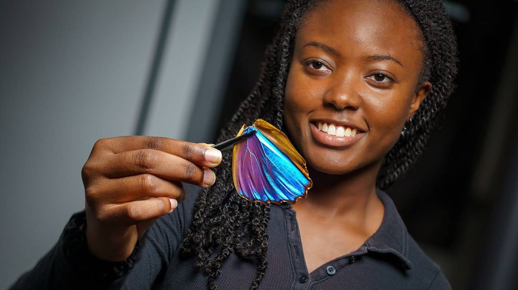 Young woman with braids smiling holds up a Morpho butterfly