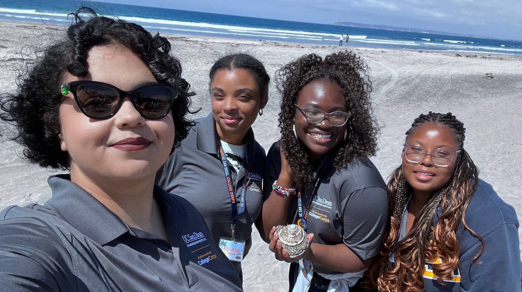Four women in gray UC San Diego College Corps polo shirts take a selfie on the beach, smiling