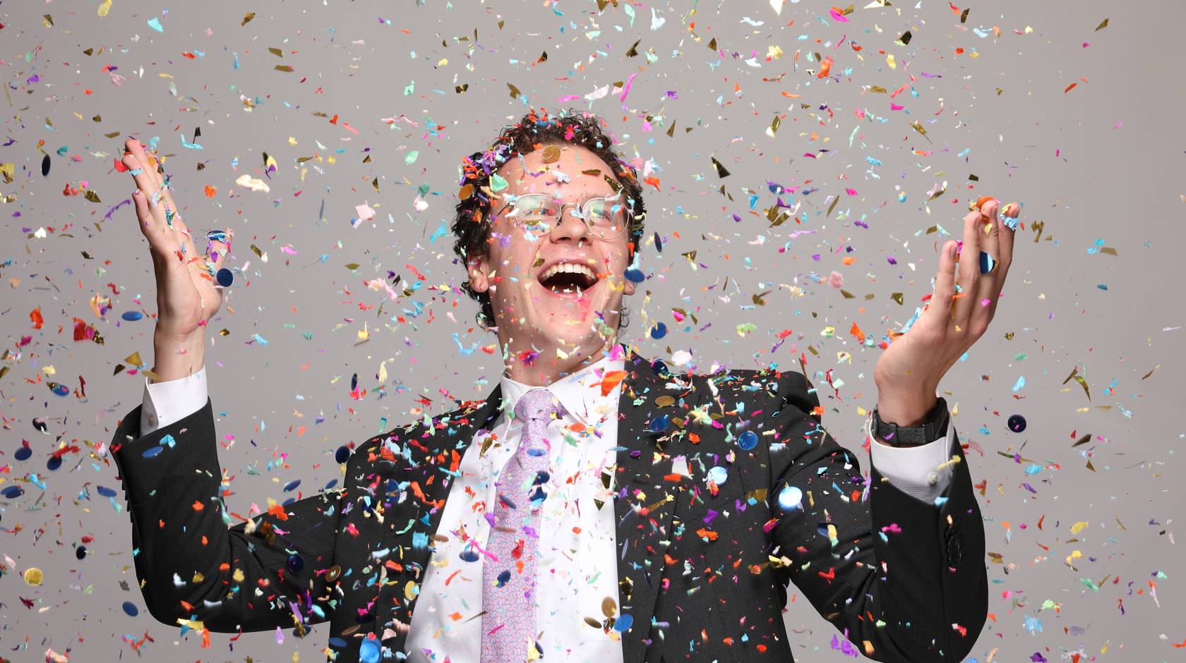An ecstatically smiling man with glasses in a suit with a pink tie holds up his arms while confetti falls