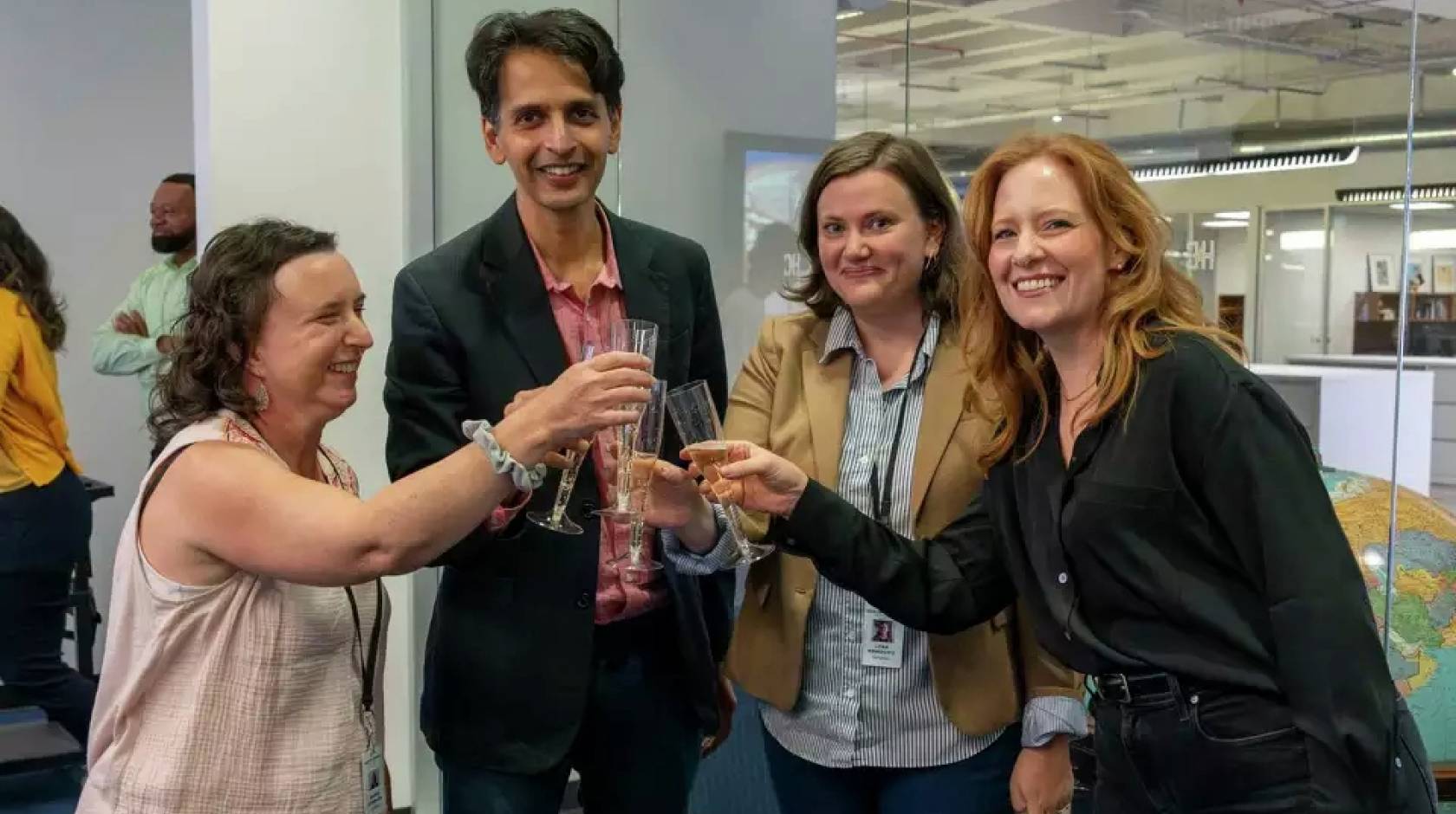 Four people in a newsroom do a champagne toast, smiling at the camera