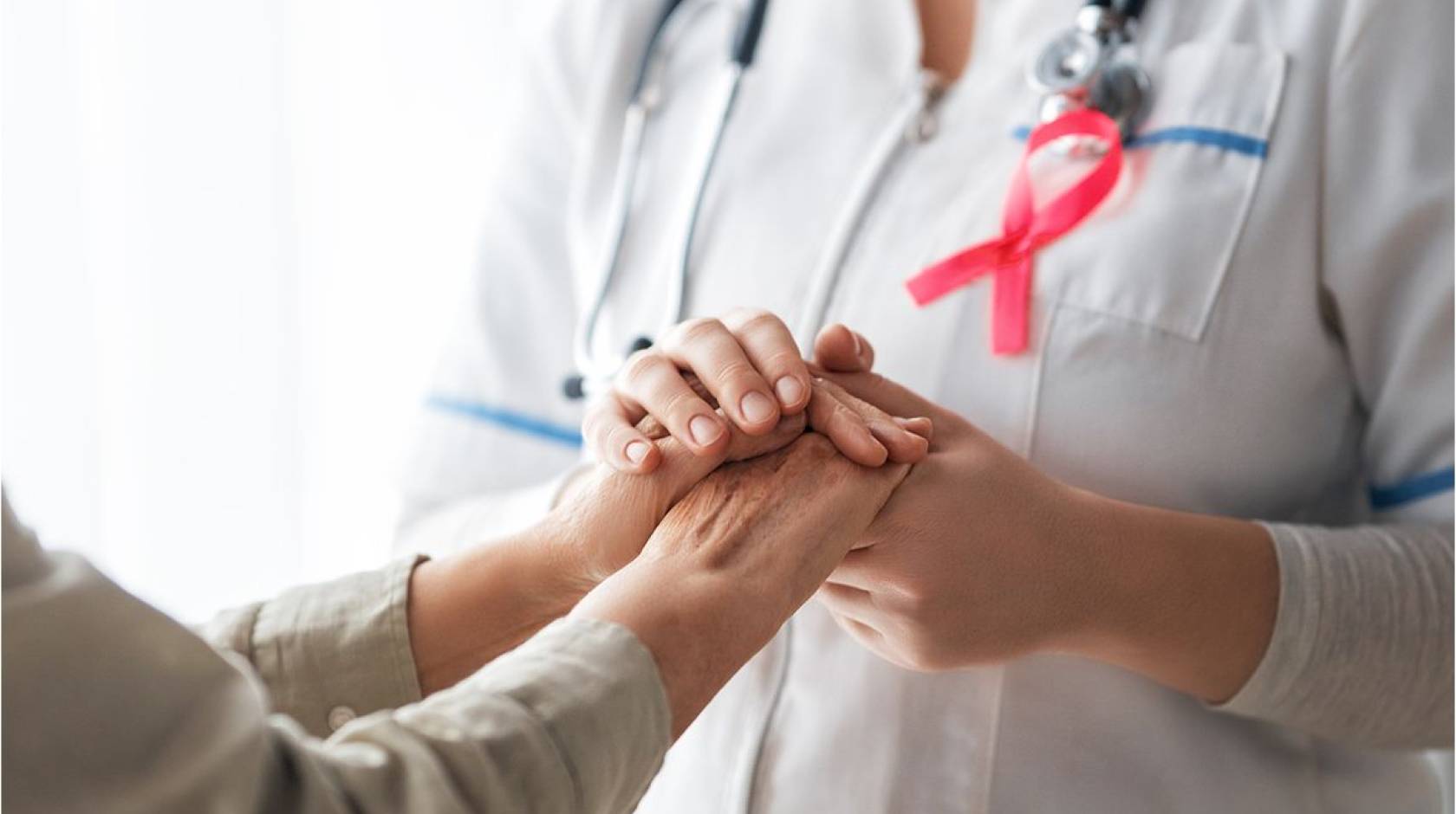 A patient, left, and medical care provider holding hands; provider has a pink breast cancer ribbon on her lapel