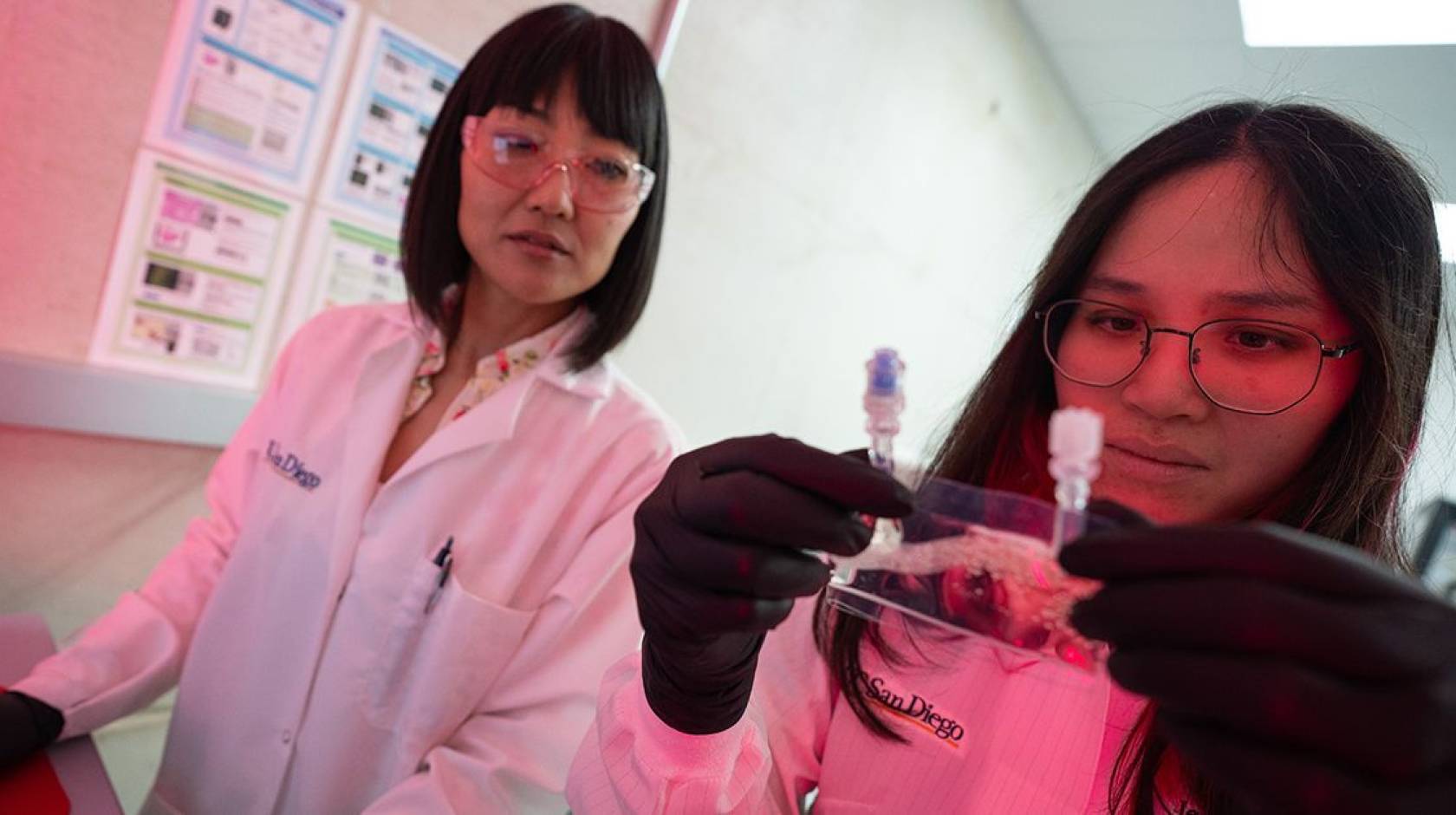 Two women in lab coats in a lab with reddish lighting in the corner; one holds and looks at a small baggie