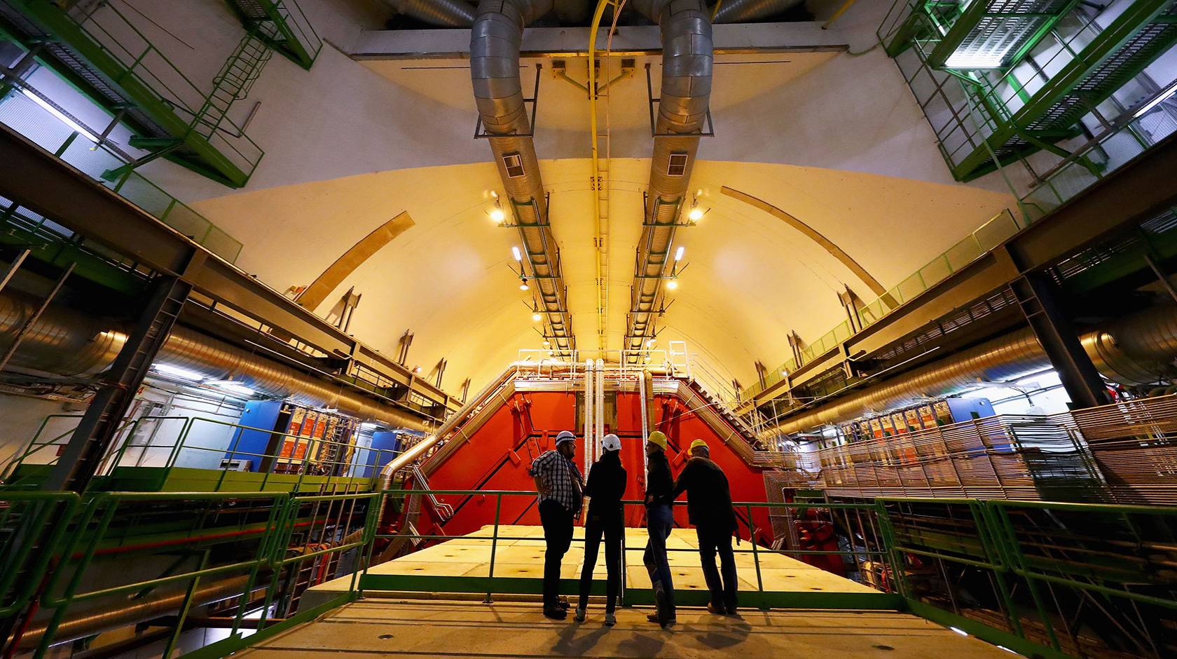 Four people stand backlit in a giant room full of high-tech machinery