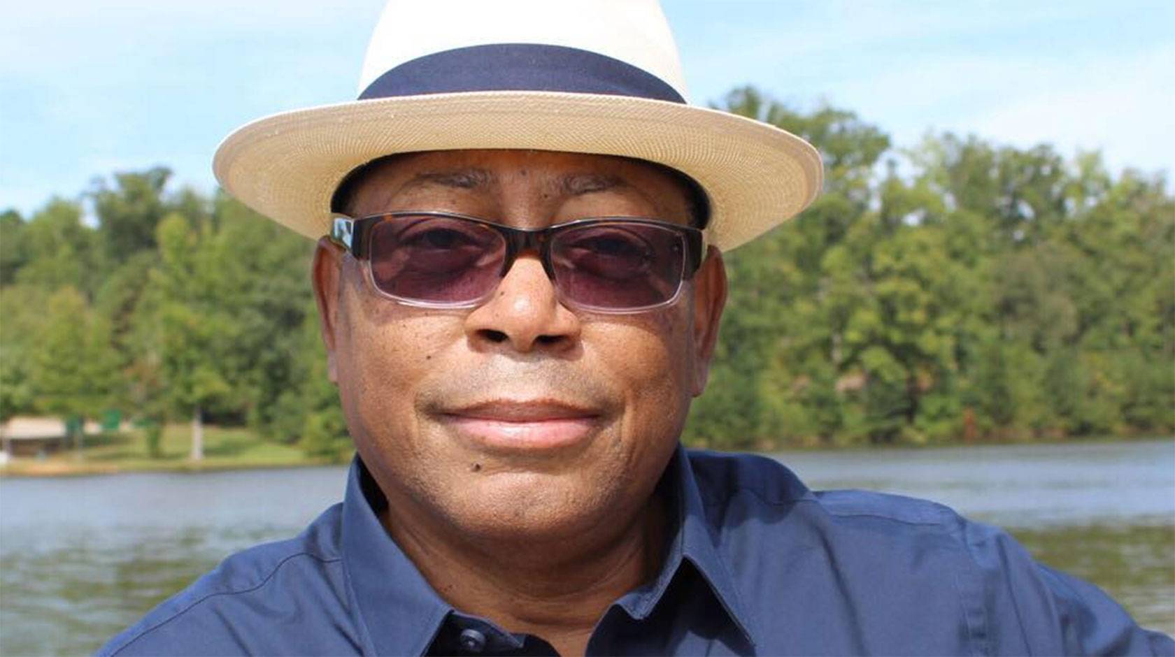 Mel Mann, wearing sunglasses and a fedora, smiles faintly, standing on the shore of a lake