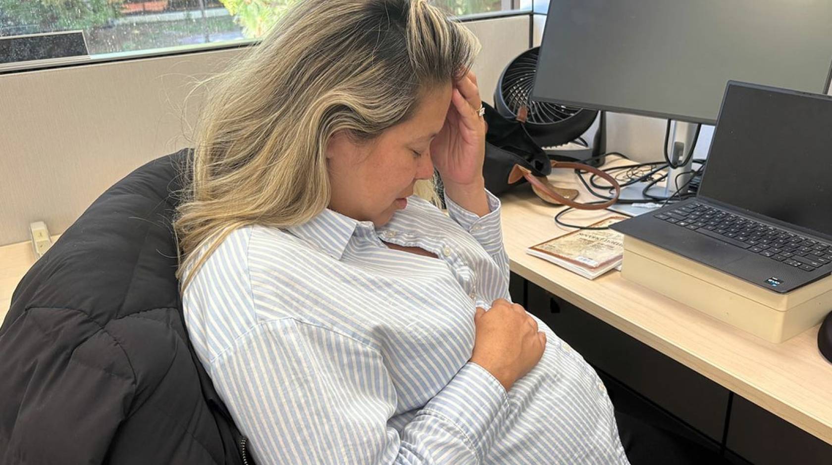 A woman holds her head with another hand over her pregnant belly while sitting at a desk in an office