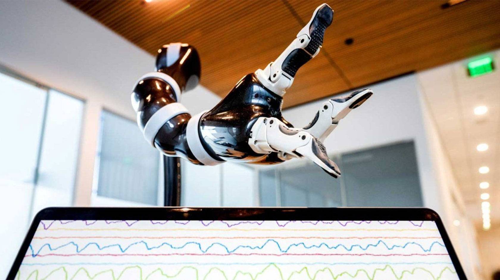 A robotic arm hovers over a computer screen showing brainwaves as a stack of tiny lines. 