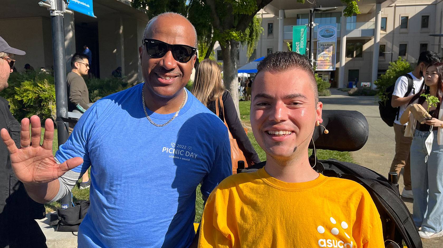 Ryan Manriquez, who uses a motorized wheelchair and wears a yellow shirt with the logo of the Associated Students of UC Davis on it, smiles for a photo alongside UC Davis Chancellor Gary May, who wears sunglasses and a blue t-shirt and is waving at the camera