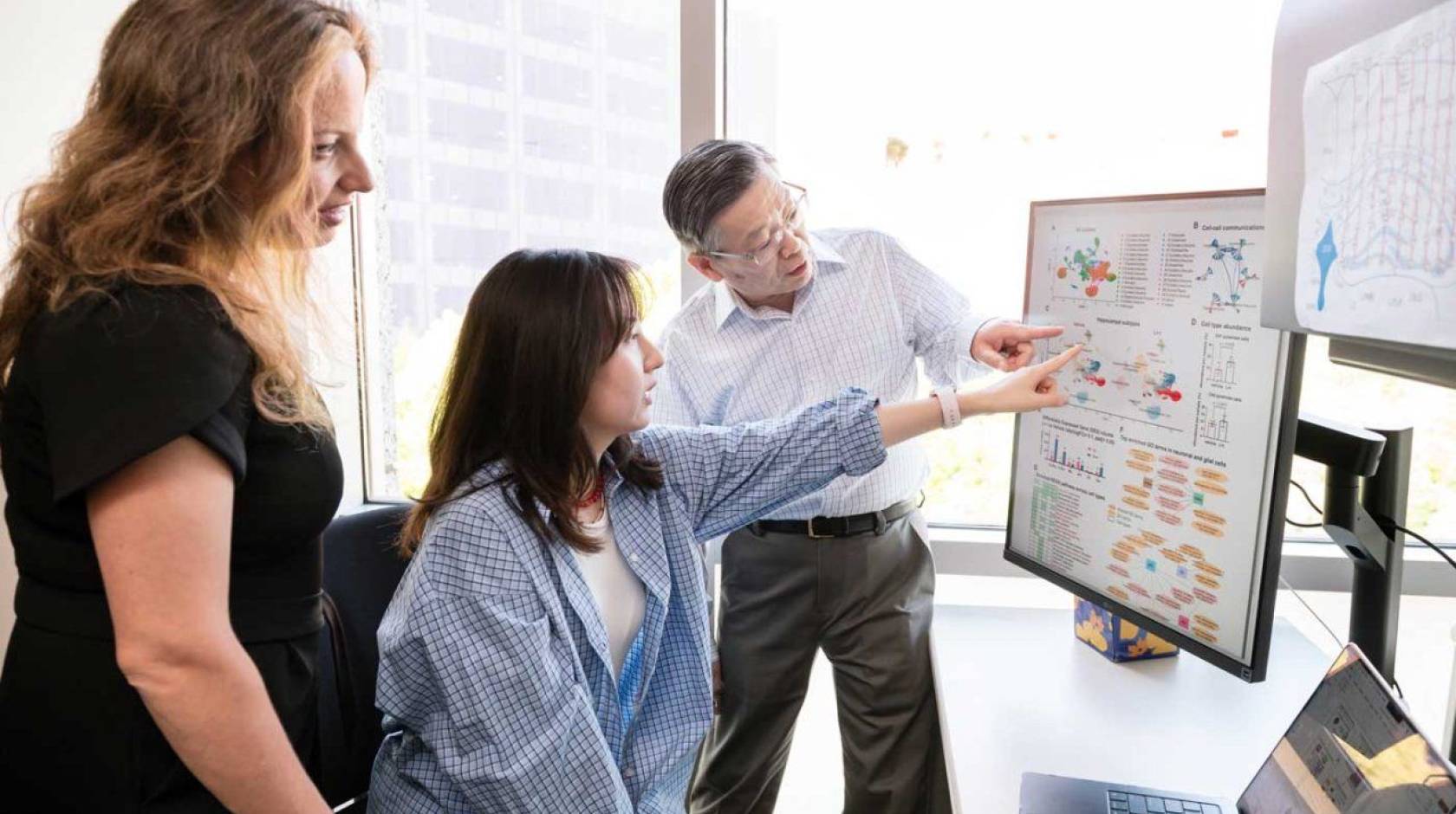 Three people in an office look at a large screen next to another large screen