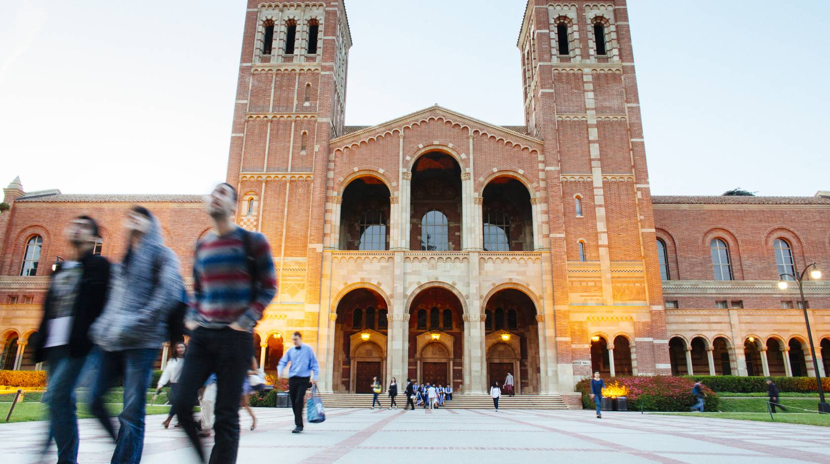Students walk in front of a large building with two towers, Royce Hall, on the UC campus as evening begins to fall