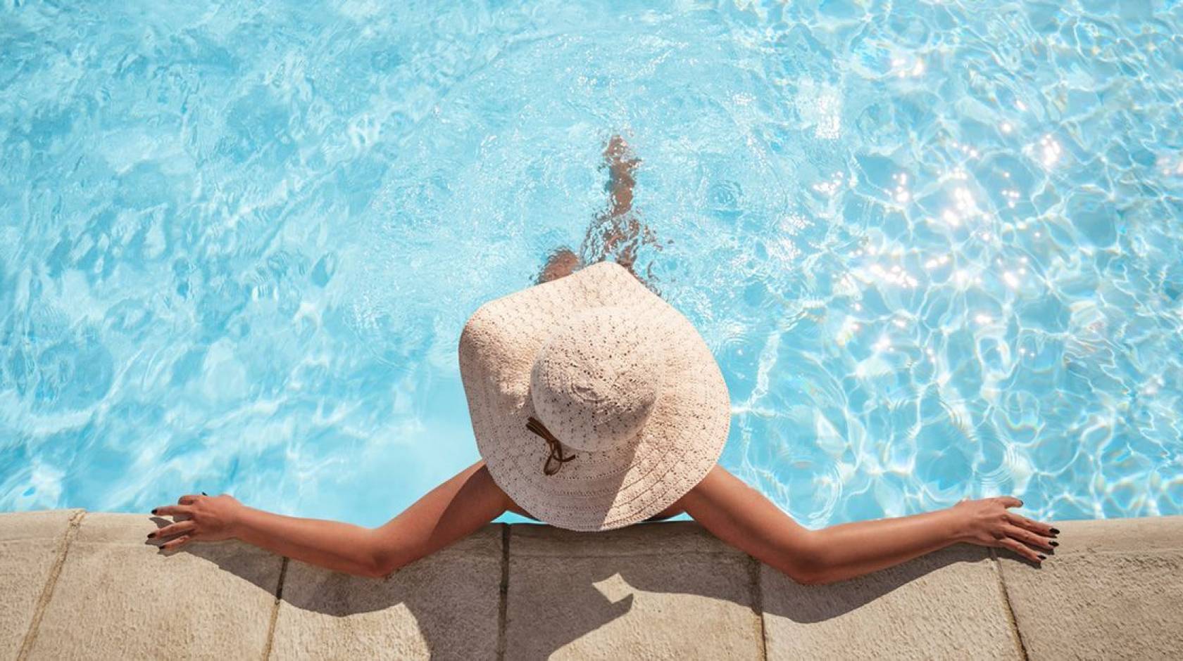 A woman wearing a brimmed straw hat relaxes at the edge of a swimming pool, photographed from above