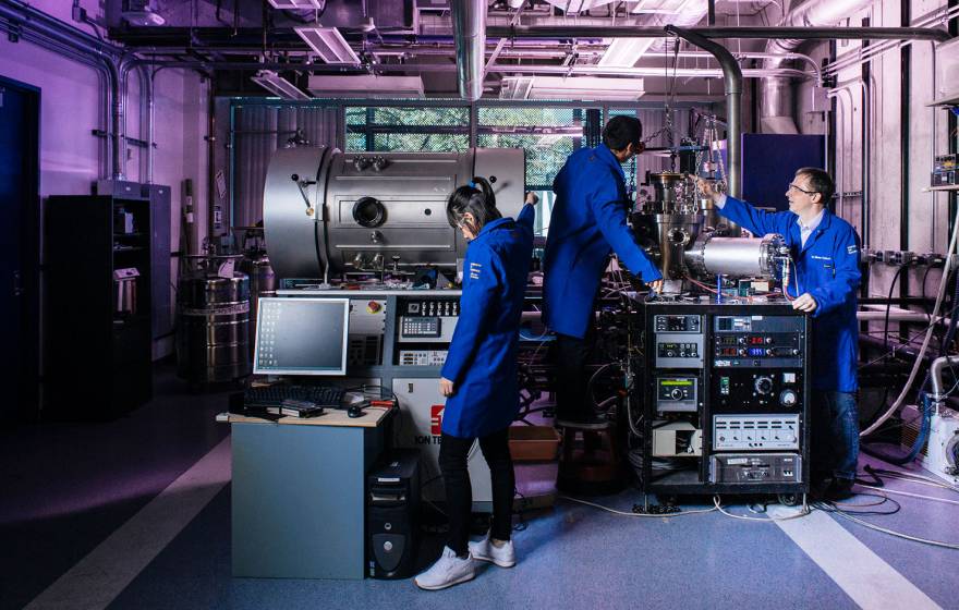 Three people in blue lab coats work in a purple-lit facility with large, complex-looking equipment.