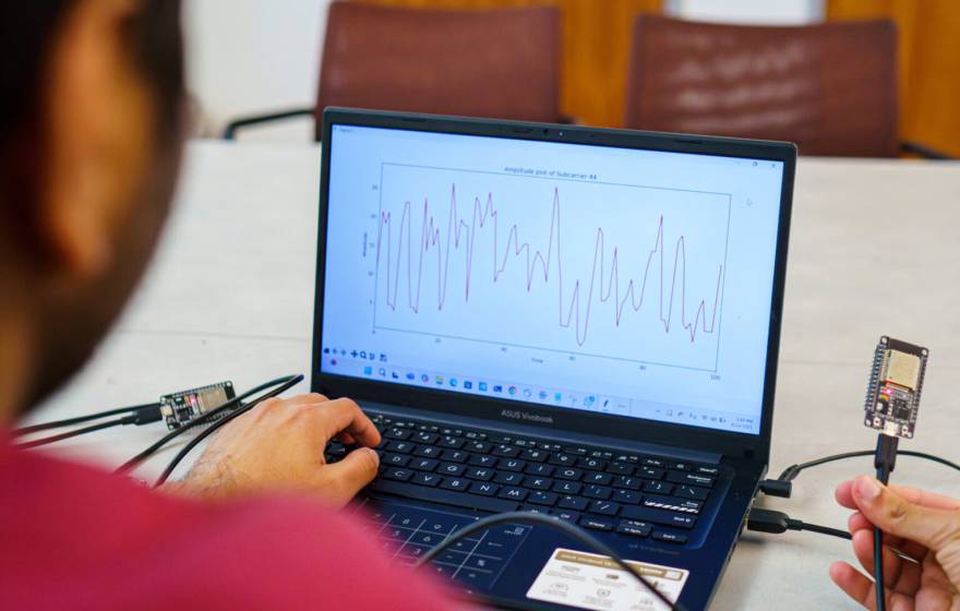 A researcher holds a wifi device in front of a laptop that displays heart rate information.