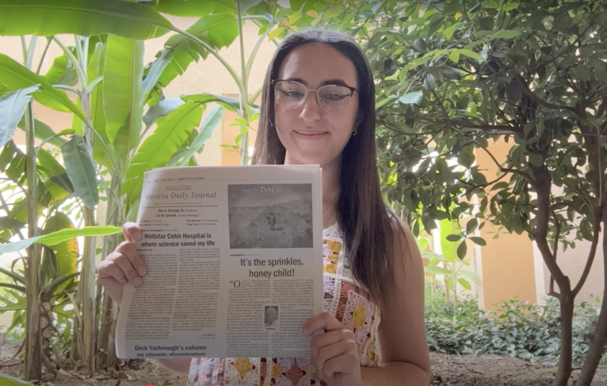 A person with long hair and glasses poses holding a newspaper