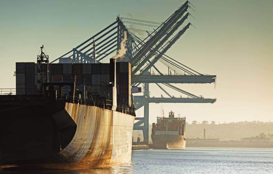 Two rusty container ships in a row, one behind, being loaded at a port during the evening