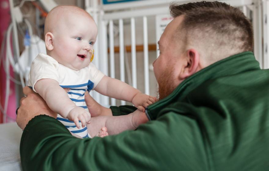 A man in a green jacket looks at a baby in front of him, both looking excitedly at each other
