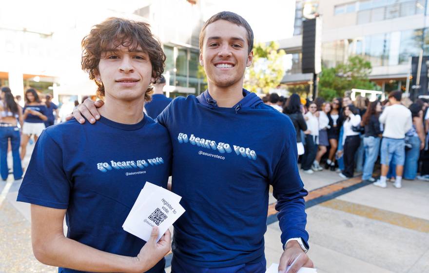 Two students smile for the camera with arms around each other's shoulders, wearing matching t-shirts that read "Go Bears Go Vote." One student holds a stack of printed QR codes labeled "Register to Vote"
