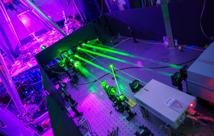 A laboratory setup with multiple bright green laser beams projected across an optical table lined with mirrors and instruments, illuminated by purple and blue ambient light, showing advanced experimental physics equipment.