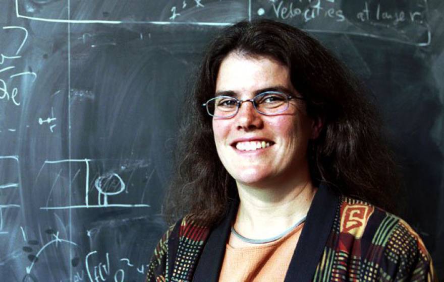 A woman wearing glasses standing in front of a chalkboard covered in scientific notations