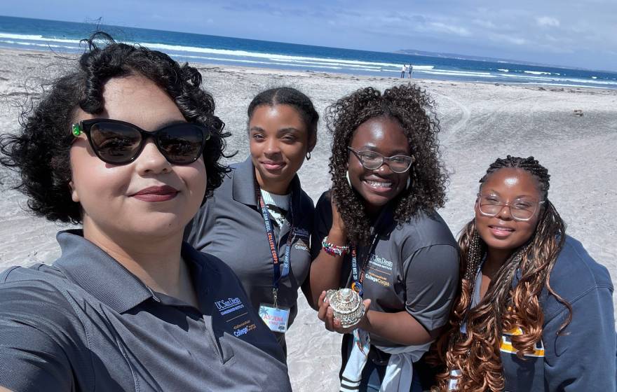 Four women in gray UC San Diego College Corps polo shirts take a selfie on the beach, smiling