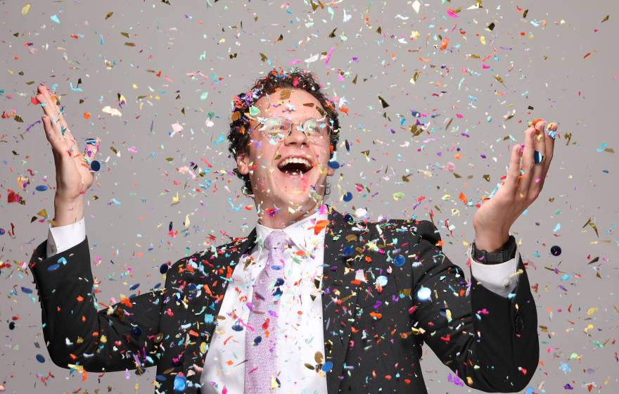 An ecstatically smiling man with glasses in a suit with a pink tie holds up his arms while confetti falls