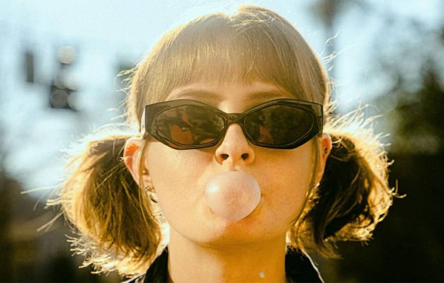 A woman wearing sunglasses and pigtails blows a gum bubble facing the camera