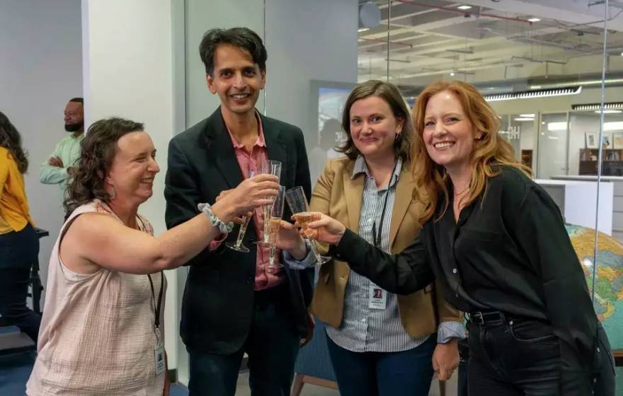 Four people in a newsroom do a champagne toast, smiling at the camera