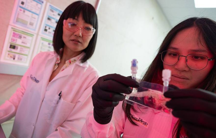 Two women in lab coats in a lab with reddish lighting in the corner; one holds and looks at a small baggie