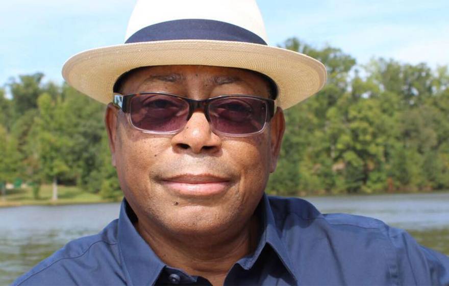 Mel Mann, wearing sunglasses and a fedora, smiles faintly, standing on the shore of a lake