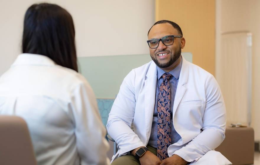 Chris Hall wearing a white medical provider's coat speaks with a patient in a lobby