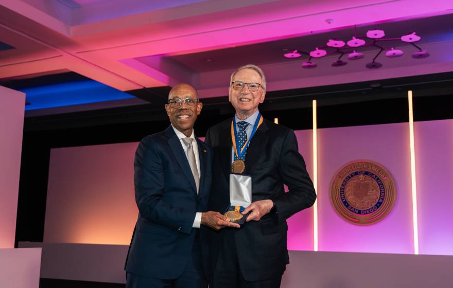 President Drake and Irwin Jacobs pose together in a room lit with purple lighting while Jacobs wears a medal and holds another
