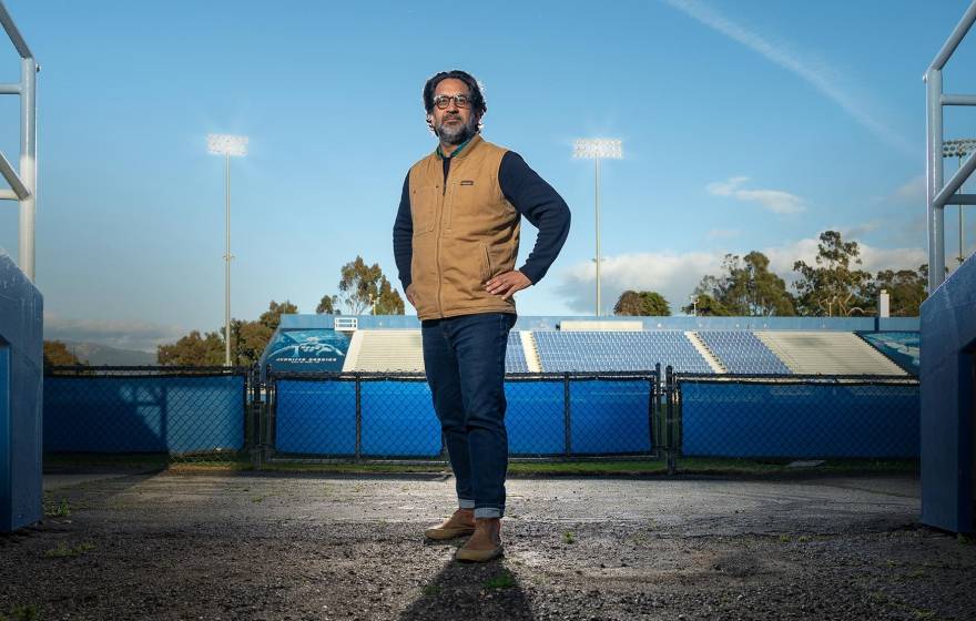 Man with salt and pepper beard stands at ground-level field entrance of a stadium, field behind him