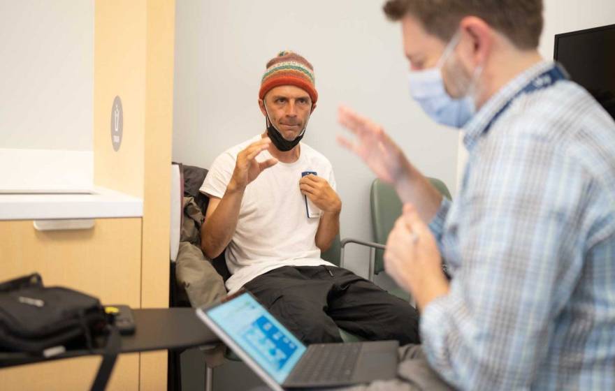 In the background, in focus, a man in a beanie makes a gesture with his hand as if to grasp. In the foreground, a doctor in a mask in front of a computer screen makes the same gesture