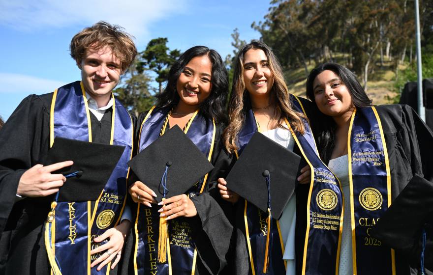 Four graduates in robes and stoles hold their caps and smile at the camera