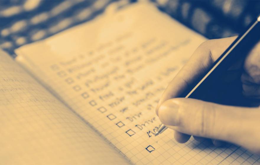 A sepia-toned close-up of a hand writing a to-do list with check-off boxes