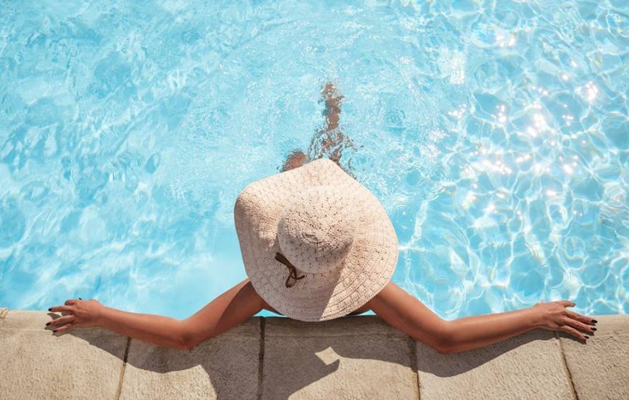 A woman wearing a brimmed straw hat relaxes at the edge of a swimming pool, photographed from above