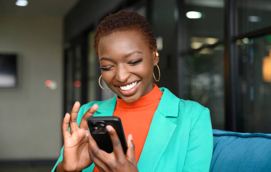 Waist-up view of shorthaired Black woman wearing hoop earrings, teal jacket over orange top, and smiling while scrolling portable device in modern office