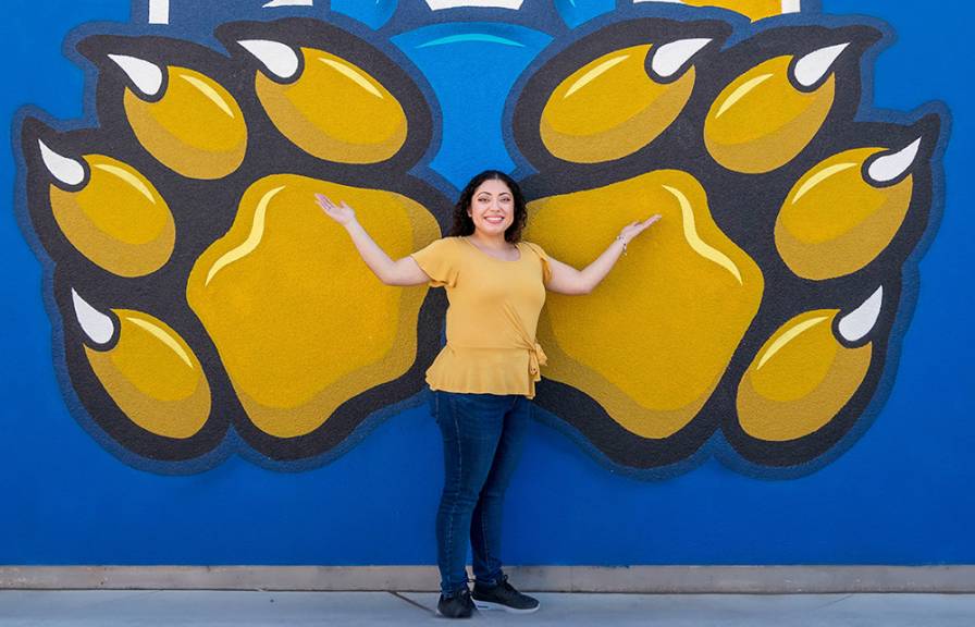 A smiling woman standing in front of a blue wall painted with the gold UC Merced bobcat paws