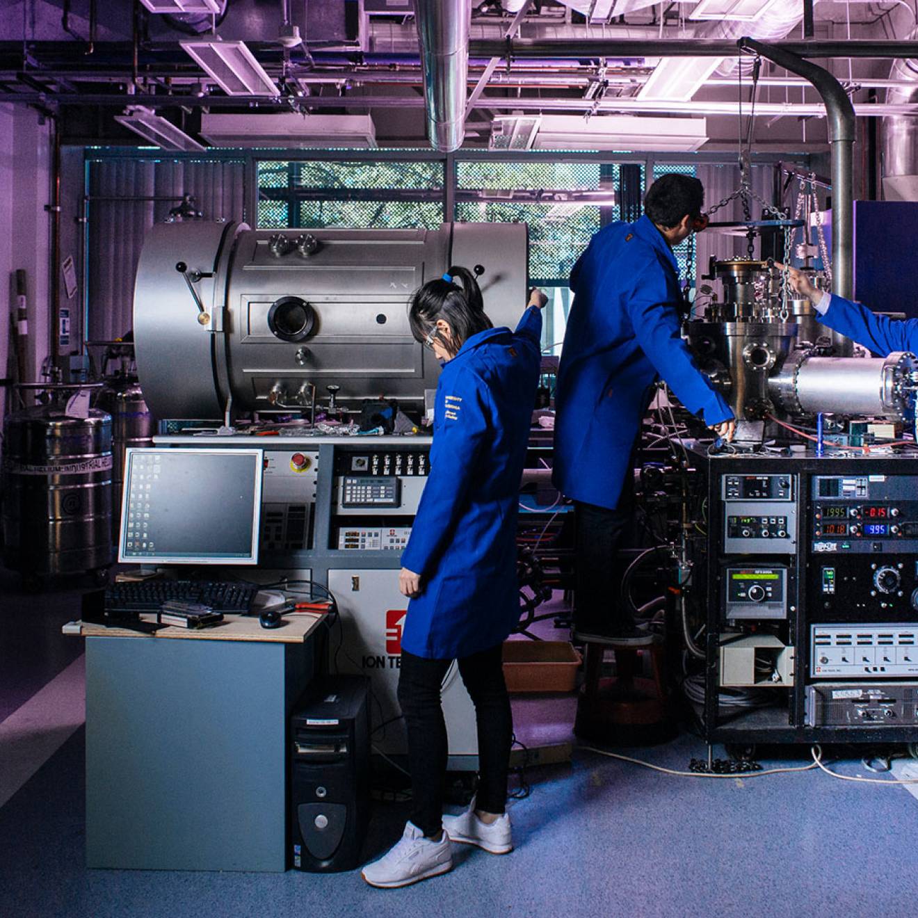 Three people in blue lab coats work in a purple-lit facility with large, complex-looking equipment.